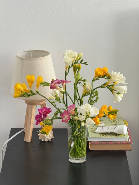 A clear glass vase containing a fresh bouquet of sweet peas in shades of white, yellow, and pink, with green stems and leaves, placed on a dark table surface. Behind the bouquet, there is a small beig