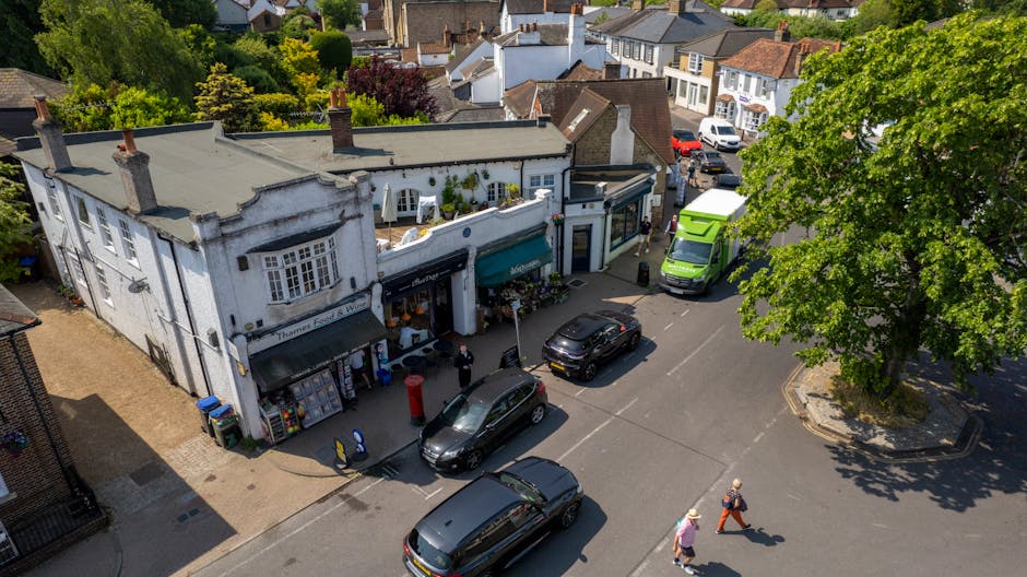 An aerial view of a small shopping street in Brockley SE4, showcasing a white, vintage-style florist shop with a blue awning and flower displays outside. The shop is surrounded by parked cars, includi