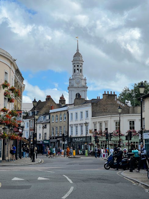 The image depicts a lively urban scene in Brockley, with a prominent historic clock tower featuring a tall spire topped by a weather vane, set against a partly cloudy sky. Surrounding the tower are ch