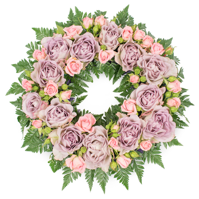 A lush, circular memorial wreath is photographed directly from above, set against a clean white backdrop that emphasises its colours and textures. The design is opulent yet serene, with the entire ring densely composed of roses and greenery. At intervals around the circle sit prominent, fully bloomed roses in soothing tones of dusty lavender, antique mauve, and soft greyish purple. Their petals form intricate spirals, some slightly cupped, others more open and flattened, creating a rich play of light and shadow within each blossom. Threaded between these larger focal roses are clusters of smaller blush pink spray roses and neat, unopened buds, whose lighter tones soften the arrangement and add a sense of youthful gentleness. The variation in rose size and stage of bloom gives the wreath a living, breathing quality, as though the flowers might still be slowly opening while it rests before a service at a Brockley chapel or a farewell gathering near Telegraph Hill. All of the blooms sit upon a substantial backing of dark green fern foliage, likely leatherleaf or a similar variety, with each frond arching gracefully outward. The finely serrated fern leaves overlap to create a dense, velvety base, peeking through between the roses and edging the entire circumference of the wreath with deep green. Small hints of darker, almost shadowy filler greens and tiny buds add depth and complexity, preventing the design from appearing flat despite its overhead presentation. The overall lighting is soft and diffused, highlighting the cool purples, gentle pinks, and glossy greens without harsh contrast. This thoughtfully composed wreath, much like those prepared by Brockley Flowers for local funerals and commemorations, expresses quiet sorrow, lasting love, and deep respect for a life remembered.